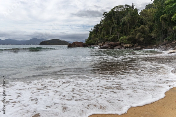 Fototapeta Ubatuba beach in São Paulo, with trees at the background, sand and waves.
Incredible paradisiac beach with cloudy sky.