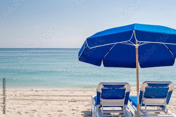 Fototapeta Two blue beach lounge chairs under a blue umbrella facing the ocean on a clear blue day