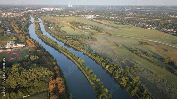 Fototapeta Aerial panoramic view of a river meandering through green fields and rural landscapes in Germany during sunrise, captured by drone.