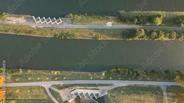 Fototapeta Aerial view of a canal with parallel paths, green vegetation and water obstacles, taken with a drone in warm morning light.