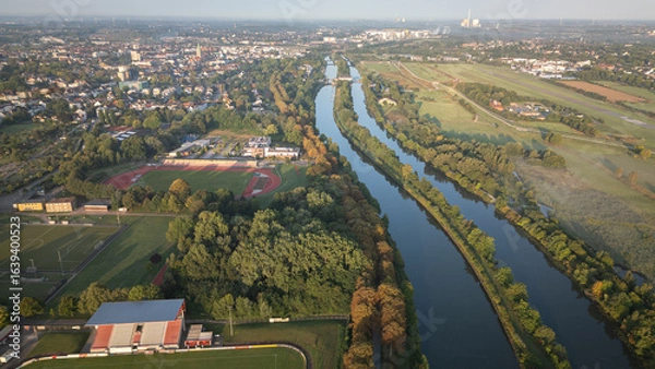 Fototapeta Aerial panoramic view of a river meandering through green fields and rural landscapes in Germany during sunrise, captured by drone.