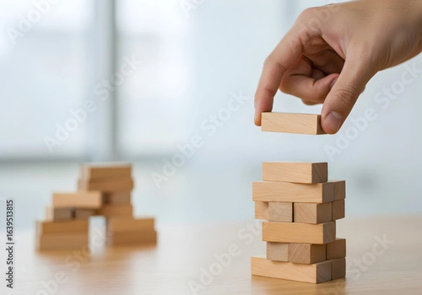 Fototapeta Building a business structure with wooden blocks. A hand carefully adds another level to a block tower, symbolizing strategic growth, project development, and building a strong foundation with precisi