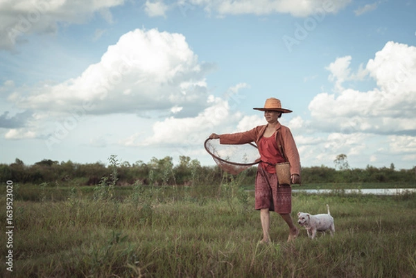 Fototapeta Serene rural landscape with farmer and dog carrying fishing net under a blue sky with fluffy clouds in a green field by the riverbank