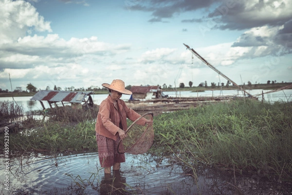 Obraz Young Fisherman in Traditional Attire Engaging in Fishing Activity on a Calm River Under a Dramatic Sky Surrounded by Nature