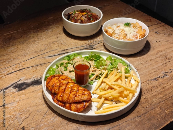 Obraz BBQ Chicken Steak - Grilled chicken breast with French Fries, fresh salad, and sauce served on a white plate over a wooden table.