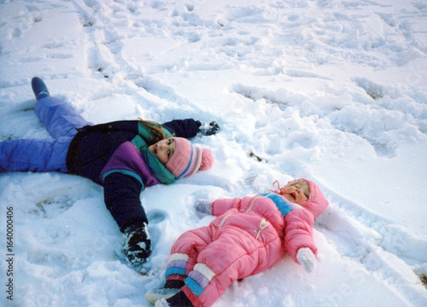 Fototapeta Children in the 80s, playing in the snow, making snow angels, wearing a snowsuit. Vintage scan, may have imperfections
