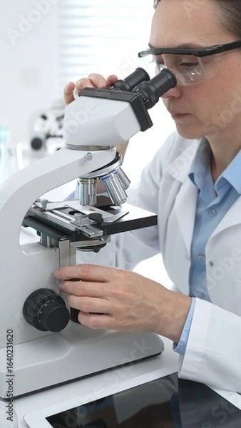 Fototapeta Female scientist wearing protective gear analyzing test samples under microscope in contemporary research environment. Medicine, healthcare and science concept