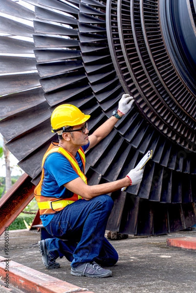Fototapeta Young Asian worker, power plant engineer in vest and helmet checking of a steam turbine, an essential component in electric power generation on outdoor with clipboard