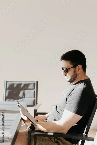 Fototapeta Man sitting on patio with sunglasses reading from an electronic tablet or book