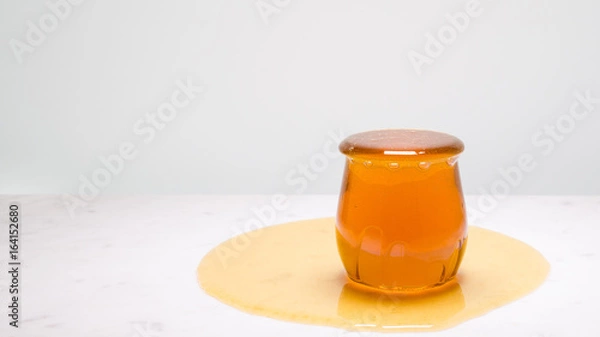 Fototapeta My cup runneth over - a jar overflowing with honey on a white marble countertop and copy space to the right.
