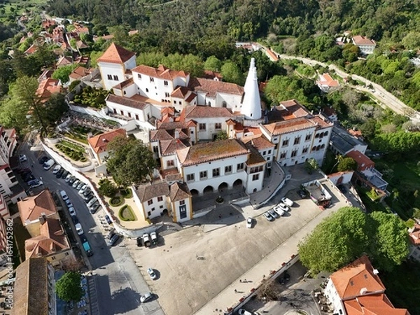 Fototapeta Aerial drone footage showing the historic town of Sintra and the National Palace of Sintra, with red rooftops, green hills, and traditional architecture captured during daylight.