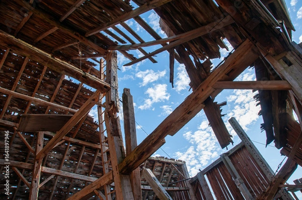 Fototapeta A unique perspective shot from inside a derelict barn, showing the dramatic collapse of the roof with a bright blue sky visible through the gaps.
