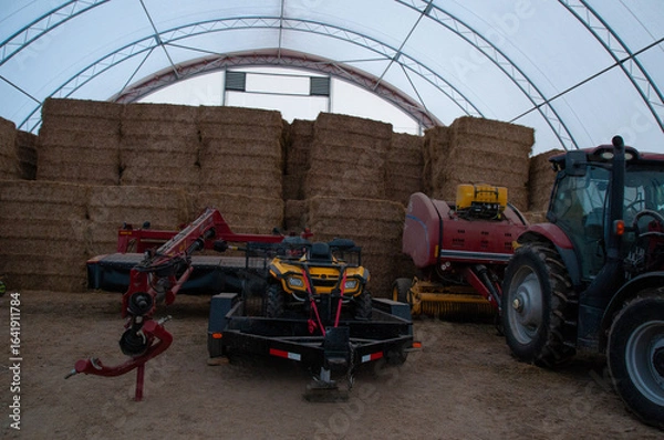 Fototapeta A wide view of various pieces of farming machinery, including a tractor, ATV, and baler, stored next to stacked hay bales inside a hoop barn.