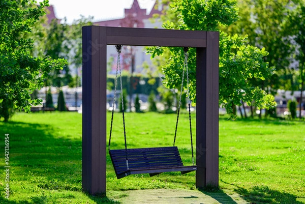 Fototapeta Wooden swing with metal chains in a public park, surrounded by green grass, trees, and sunlight on a bright summer day, peaceful outdoor recreation and relaxation scene