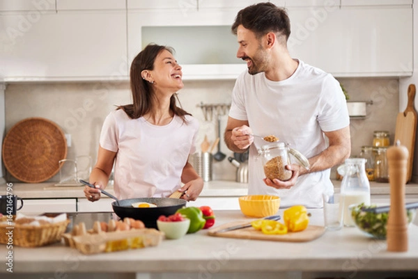 Fototapeta In a modern kitchen, a couple cooks breakfast together, smiling and sharing moments of joy. They prepare ingredients while enjoying each other's company, creating a joyful atmosphere.