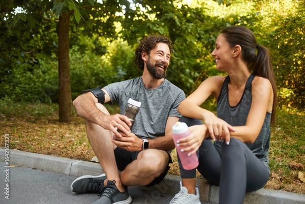 Fototapeta Two friends relax and smile while taking a break after exercising in a lush park. They are enjoying their drinks and celebrating a healthy lifestyle together.