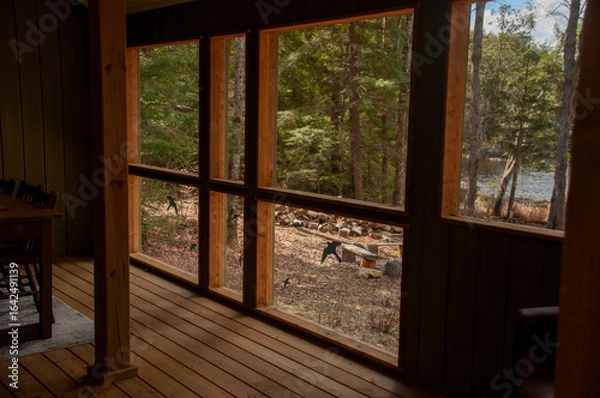 Fototapeta A cozy, rustic cottage interior with wooden floors and paneling, looking out through large windows to a wooded landscape and a sunlit lake.