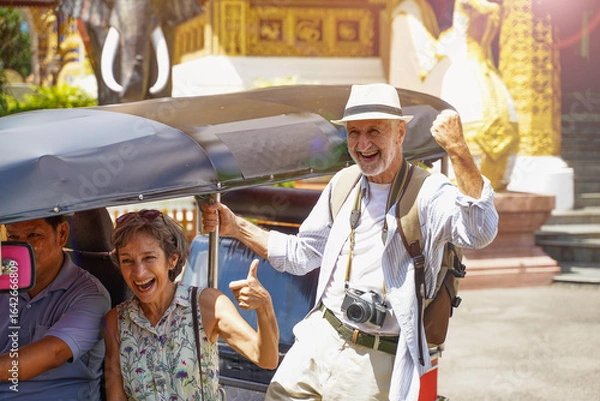 Fototapeta Senior American tourist couple showing funny smiles and laughter while taking photos with Tuk Tuk Thailand taxi in a Thai temple area with sun bright background. Senior tourist concept