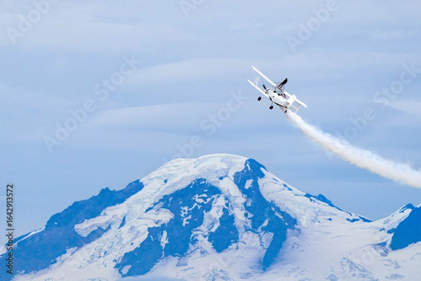 Obraz Wing walker on a vintage biplane climbs above a snow-capped mountain, white smoke streaming across a blue sky, a dramatic aerobatics symbolizing courage, freedom and high altitude adventure.