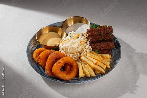 Fototapeta A rich beer snack platter with crispy onion rings, golden French fries, cheese strings, toasted rye bread with dill and two sauces served on a blue ceramic plate