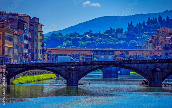 Fototapeta Santa Trinita bridge with three arches spans the Arno River, flanked by historic buildings. In the background. Tree-lined hills and structures rise in the distance under a clear sky. Florence, Italy.