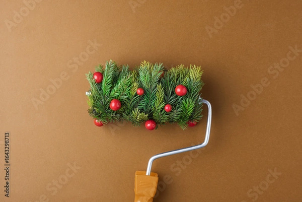 Fototapeta Festive paint roller with fir sprigs and red berries on warm brown backdrop promises cozy holiday makeover and playful Christmas mood.