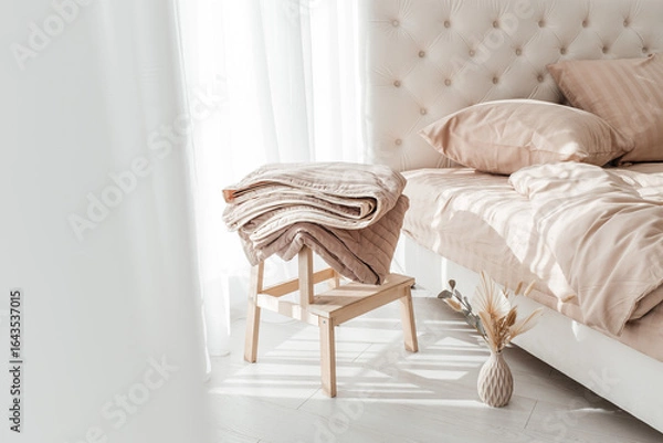 Fototapeta Cozy folded beige blanket on wooden stool next to a neatly made bed in morning sunlight