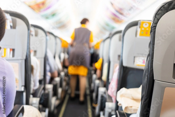 Fototapeta Inside commercial airplane, passengers sit calmly in economy class. Selective focus of the aisle, with blurred seats creating depth. It shows air travel, aviation lifestyle, or transportation scenes.