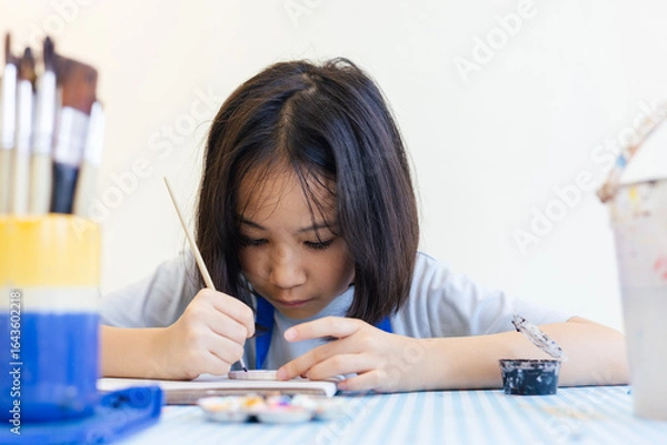 Fototapeta Young Asian girl engages deeply in a painting activity at school, surrounded by colorful paints and brushes. Her concentration reflects early artistic development in a creative learning environment.