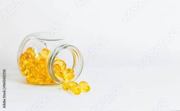 Fototapeta Glass jar with yellow vitamin capsules spilled on white surface. Clean layout with copy space emphasizes softgel fish oil supplements, wellness lifestyle, and modern healthcare concept.
