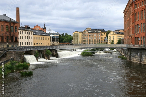 Fototapeta Photo with a view of former factory buildings, a smokestack and a bridge over a river in the center of Norrköping in Sweden