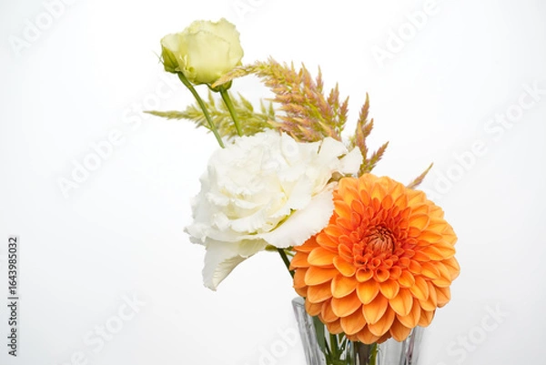 Fototapeta Small floral arrangment of orange and white flowers in a glass vase. White background. Dahlia, lisianthus, and celosia flowers.