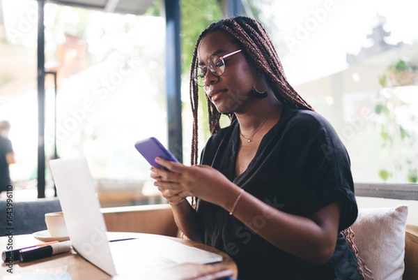 Fototapeta Concentrated young Black woman checking phone while working on laptop, immersed in digital tasks, reflecting flexibility and multitasking in mobile tech-based freelance environment.