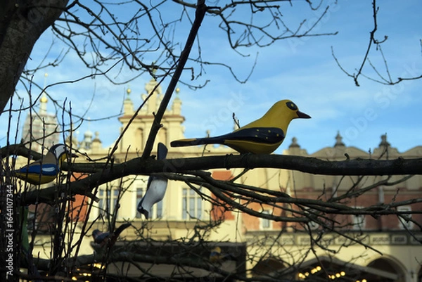 Fototapeta A leafless tree with several colorful artificial birds perched on its branches is set against a clear blue sky and urban buildings in the background.