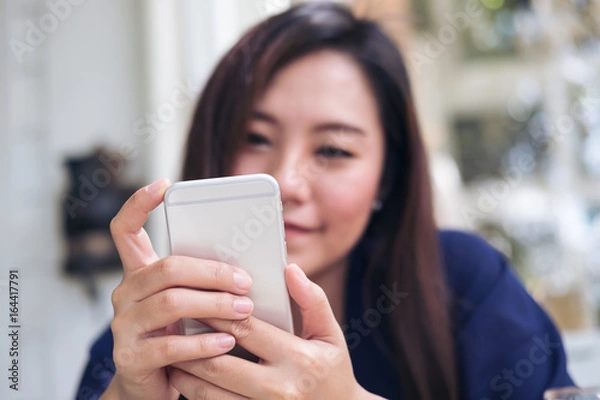 Fototapeta Beautiful Asian woman with smiley face using and looking at smart phone in modern cafe