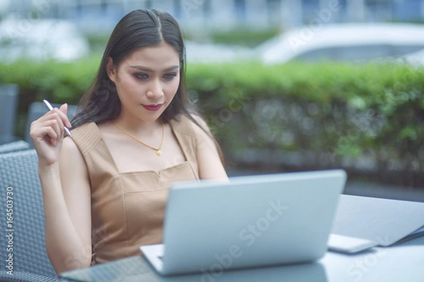 Fototapeta Sharing good business news. Attractive young woman talking on the mobile phone and smiling while sitting at her working place in office and looking at laptop, businesswoman working. Selective focus.