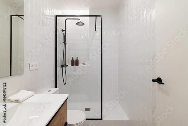 Fototapeta Modern bathroom with a glass shower enclosure, white tiles, and a wooden vanity. Minimalist design with toiletries on the shelf and a clean aesthetic.