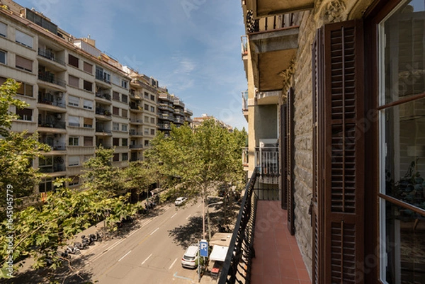 Fototapeta A sunny street view from a balcony in a city. Buildings line the street, with trees providing shade. Clear blue sky enhances the urban landscape.