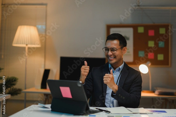 Fototapeta Asian businessman showing thumbs up during video call on tablet in office at night