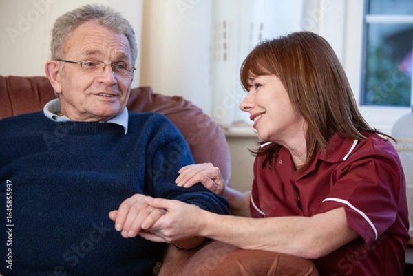 Fototapeta An elderly man visited at home by his friendly care worker who holds his hand listening intently with a smile while he talks to her.
