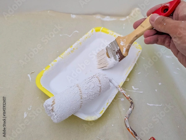 Fototapeta Painter's hand holding red-handled brush while carefully taping around electrical outlet during wall preparation process