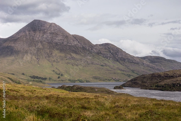 Fototapeta Assynt Peninsula, Scotland - June 7, 2012: Brown Mountain range along Loch Lurgainn under silver sky with gray clouds. Green vegetation on foothills and dry foreground.