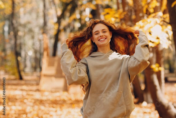 Fototapeta Smiling young woman with curly hairstyle enjoys the autumn weather in the forest with the yellow leaves at sunset