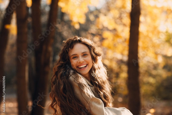 Fototapeta Smiling young woman enjoying the autumn season in the fall park with the yellow leaves at sunset