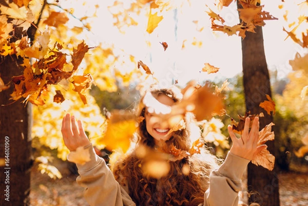 Fototapeta Smiling young woman throw up the autumn leaves in the forest at sunset