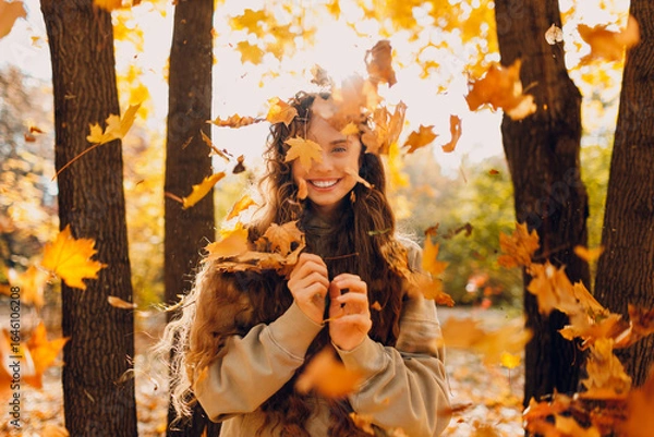 Fototapeta Smiling positive young woman throws up the yellow autumn leaves in the forest at sunset