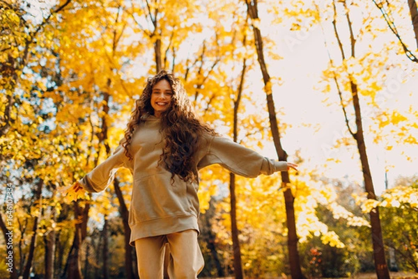 Fototapeta Smiling young woman plays and jumping dancing in the autumn forest with the yellow leaves at sunset