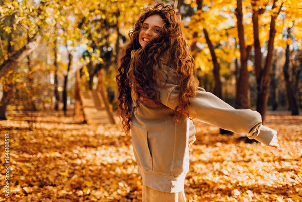 Fototapeta Smiling young woman enjoys the autumn weather in the fall park with the yellow leaves at sunset