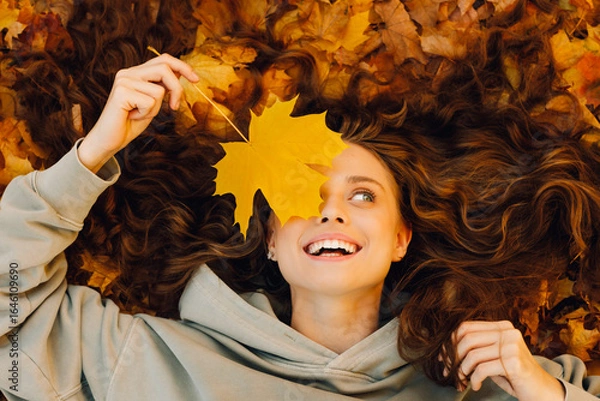 Fototapeta Smiling young woman lies on the autumn foliage in the forest with the yellow leaves