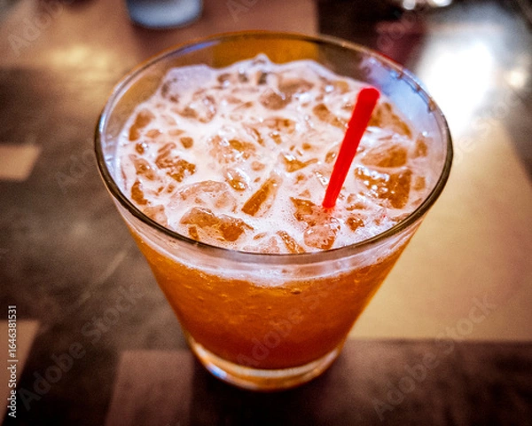 Fototapeta A classic chilled Mai tai cocktail served over crushed ice in a short glass, featuring a frothy orange hue and garnished with a red straw, set on a softly lit table.